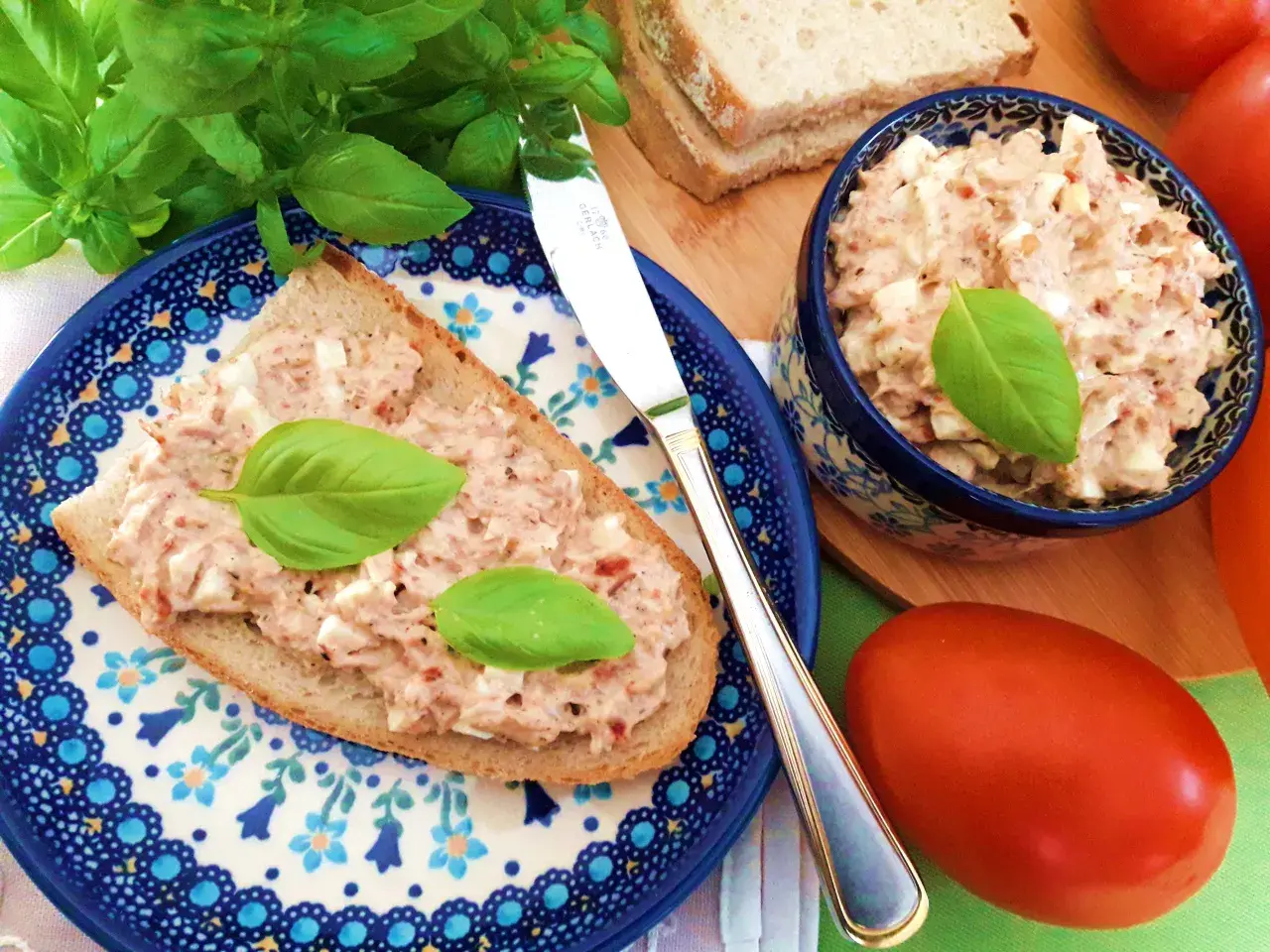 Pyszna pasta jajeczna z tuńczykiem na kromce chleba, ozdobiona bazylią. Obok miseczka z pastą i świeże pomidory.