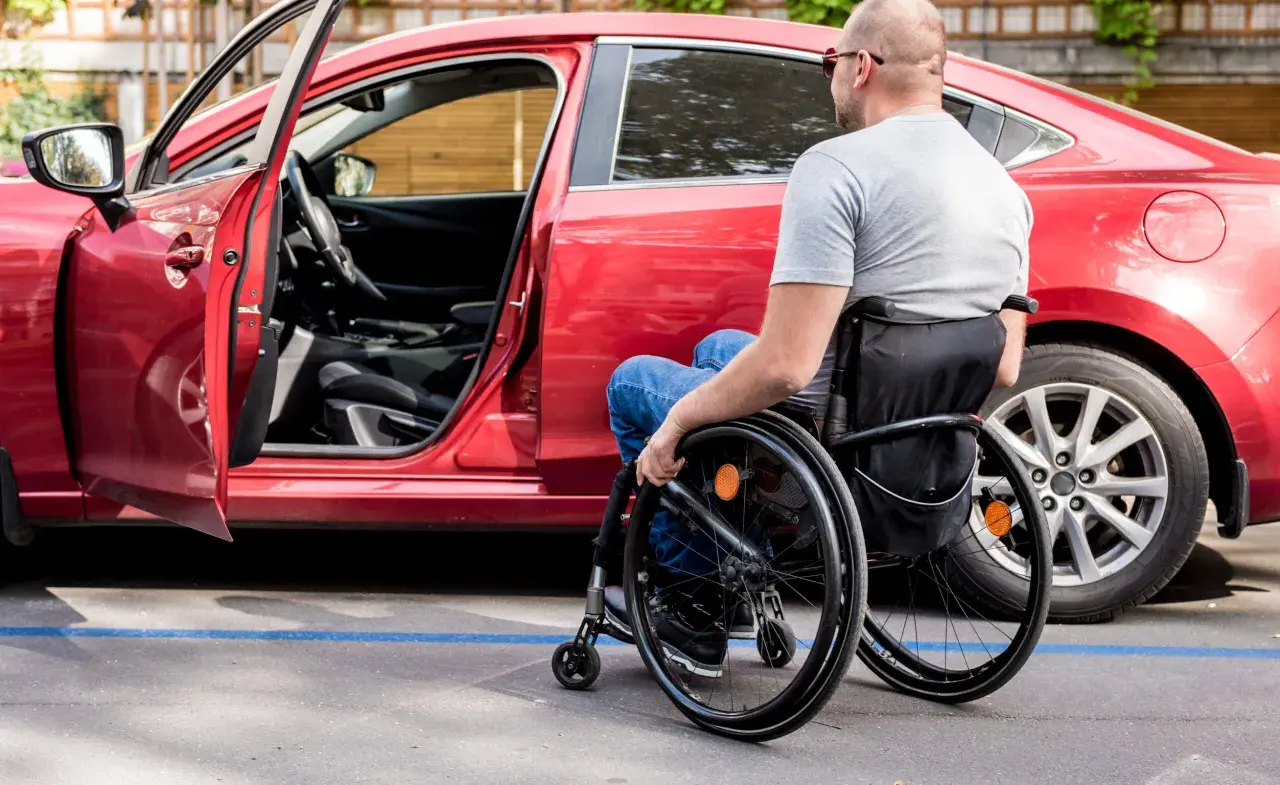 Uomo in carrozzina accanto a un'auto rossa, con portiera aperta. Un esempio di accessibilità, come previsto dall'art. 30 comma 7 legge 388 del 2000.