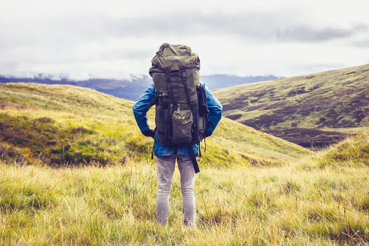 Wanderer mit großem Rucksack steht auf einem Hügel, bereit für seine 1 Jahr Weltreise. Die Landschaft ist grün und hügelig.