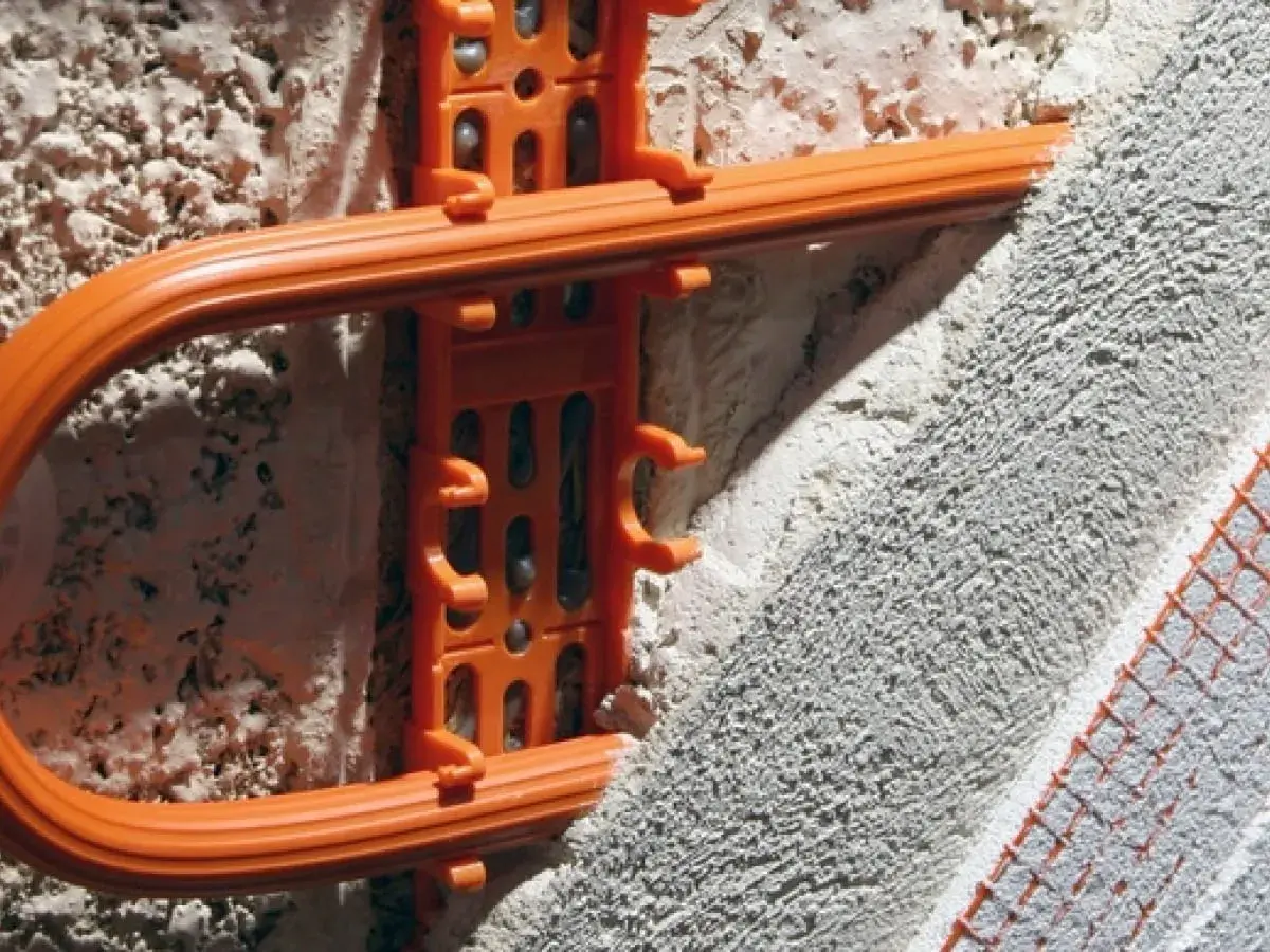Montaż ogrzewania ściennego w starym domu. Pomarańczowe uchwyty mocują rurę grzewczą do ściany, przygotowując ją do otynkowania.