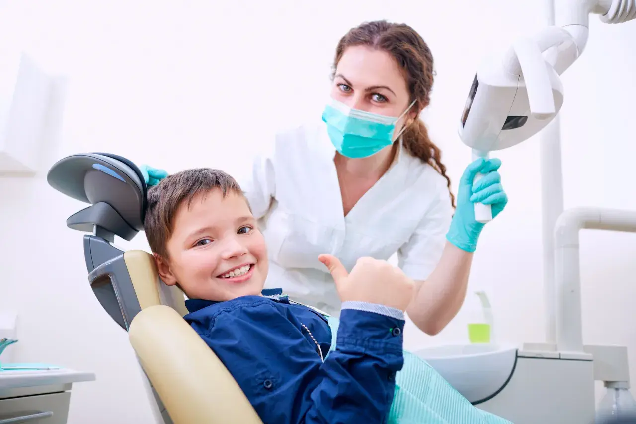 Uśmiechnięty chłopiec daje kciuk w górę w gabinecie stomatologicznym. Uśmiechnięta dentystka w masce i rękawiczkach stoi za nim.