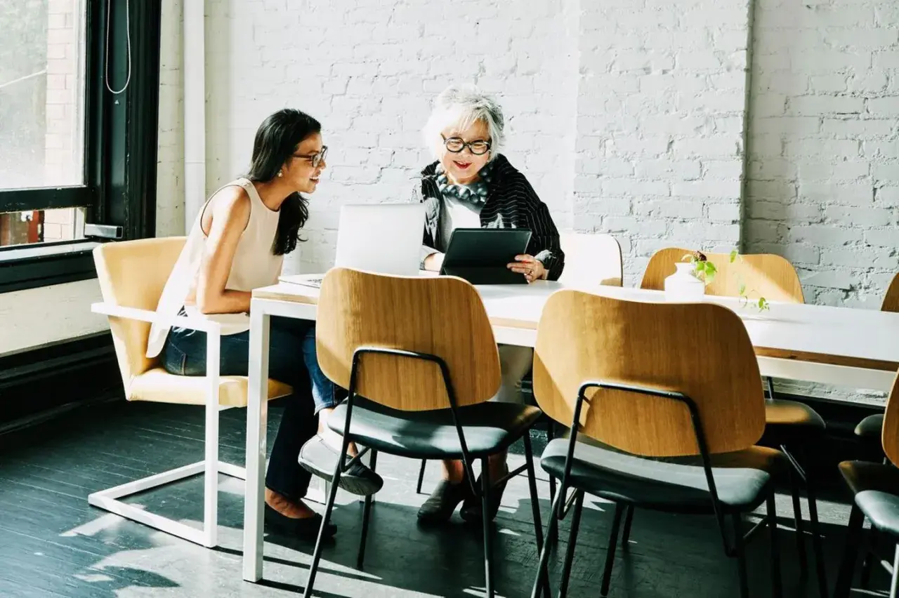 Zwei Frauen besprechen am Tisch die betriebsrente. Nachteile werden diskutiert, während sie auf Laptops und Tablets schauen.