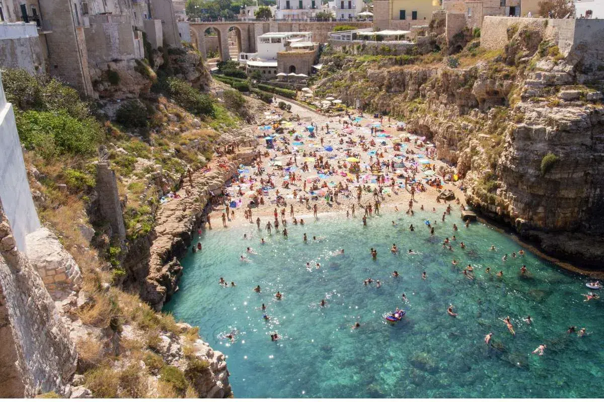 Una spiaggia affollata a Polignano a Mare, Italia, con persone che prendono il sole e nuotano nelle acque cristalline.