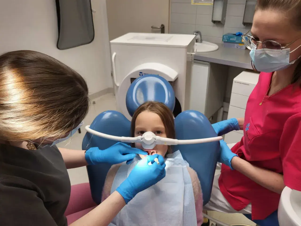 Dziecko u dentysty z maską do podawania gazu rozweselającego. Dwie higienistki dbają o komfort małego pacjenta podczas zabiegu.