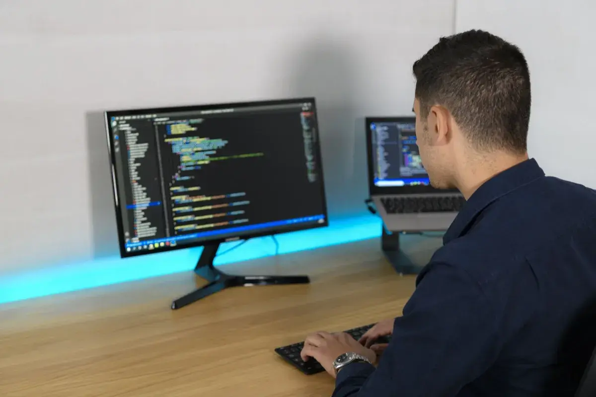 Ein Mann tippt Code auf einer Tastatur, während er auf zwei Bildschirme blickt. So macht ein Programmierer seine Arbeit.