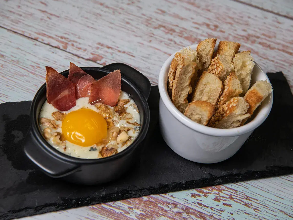 Un œuf cocotte raclette, crémeux et gourmand, garni de noix et de fines tranches de viande séchée, servi avec des toasts croustillants.