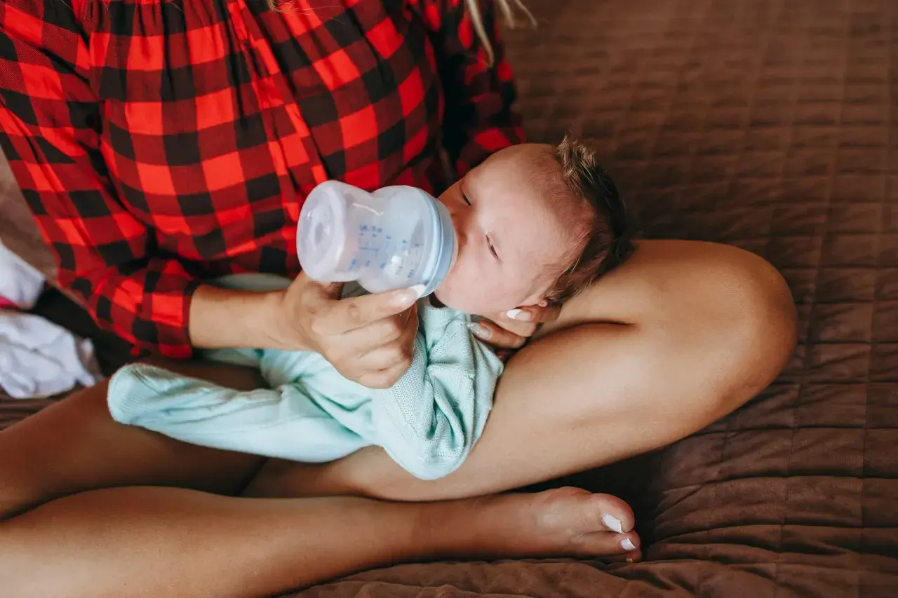 Mamma con maglia a quadri rossi offre il biberon a un neonato, praticando l'allattamento misto.
