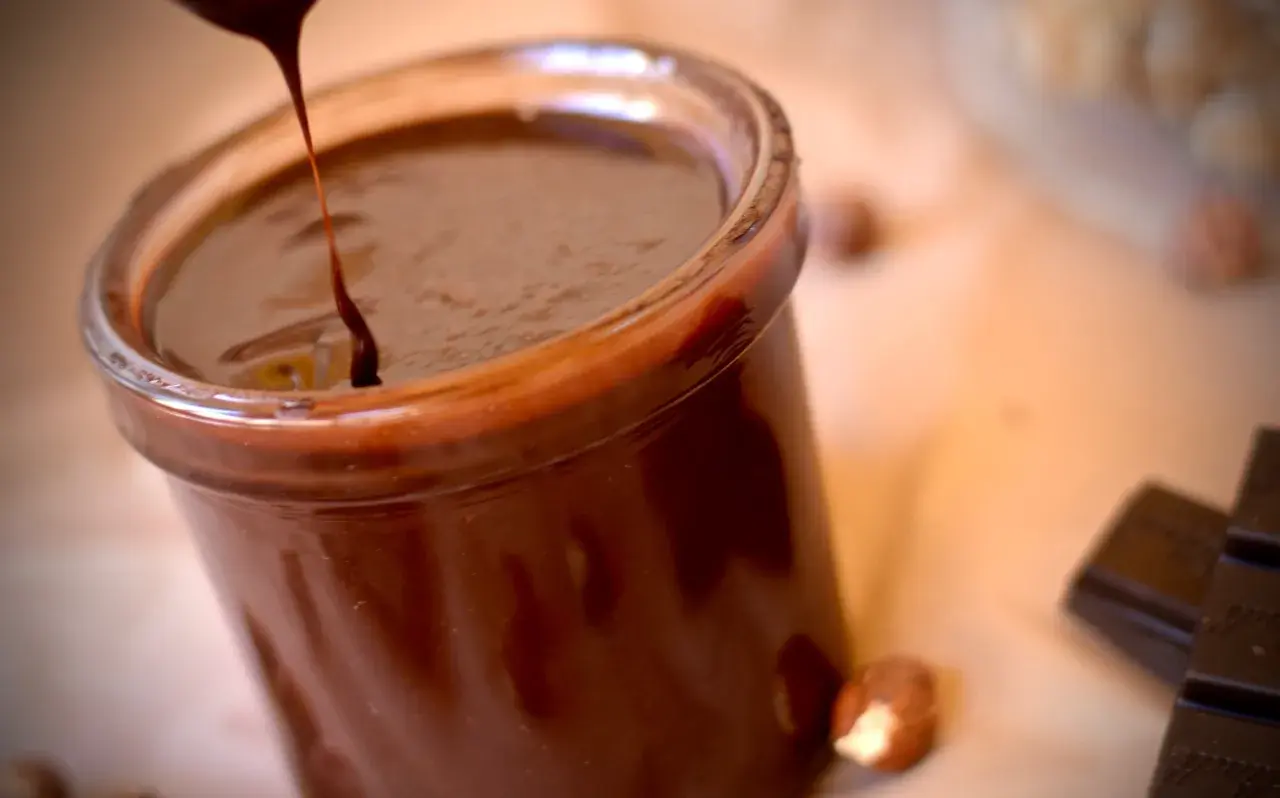 Un chorrito de crema de chocolate cae en un frasco lleno de esta delicia. Avellanas y tabletas de chocolate completan la escena.