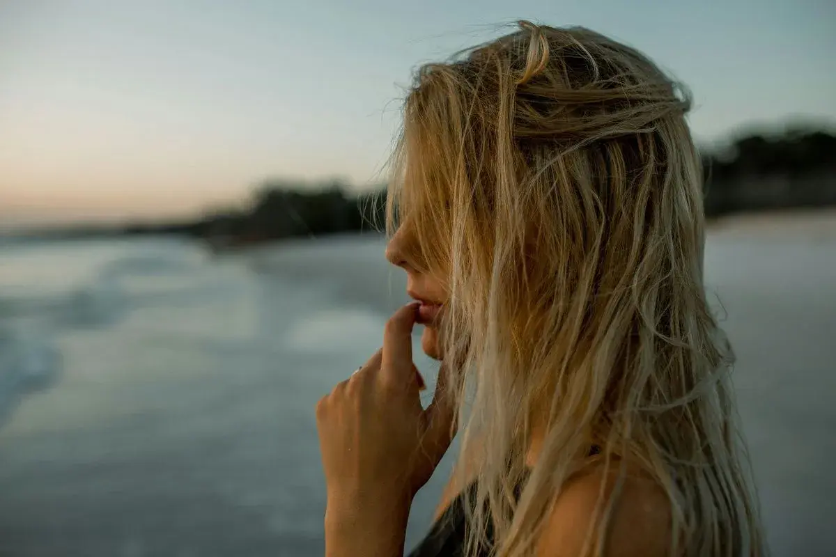 Kobieta z blond włosami rozjaśnionymi przy twarzy, na tle plaży i morza.