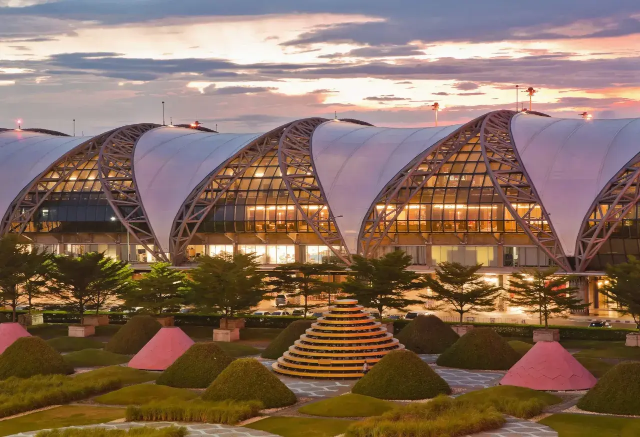 Der moderne Flughafen Bangkok Suvarnabhumi mit seiner markanten Dachkonstruktion und der kunstvollen Gartenanlage bei Sonnenuntergang.