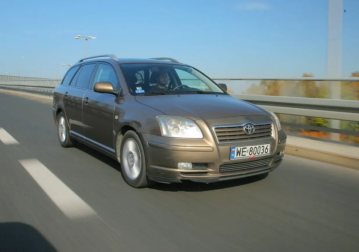 Brązowy Toyota Avensis T25 w kombi jedzie po drodze. Widać przednie żarówki i tablicę rejestracyjną WE 80036.