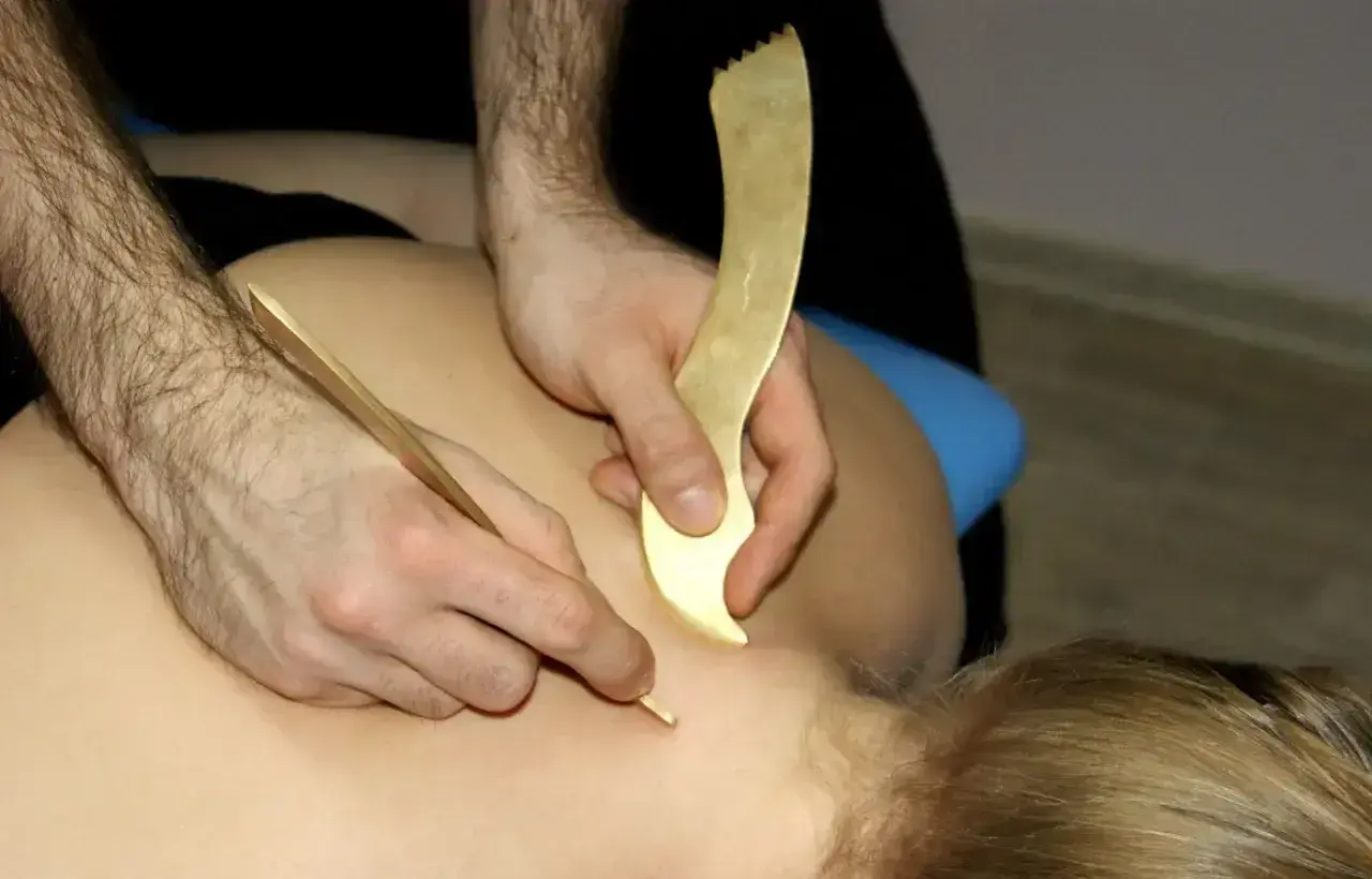 Masaż narzędziami do pinoterapii. Terapeuta pracuje nad karkiem pacjenta, używając złotych przyrządów do głębokiego masażu tkanek.