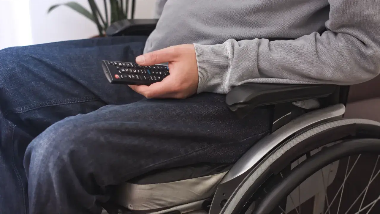 Osoba na wózku inwalidzkim, z umiarkowanym stopniem niepełnosprawności, trzyma pilot do telewizora.