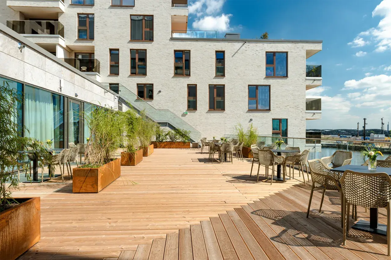 Holzterrasse mit Tischen, Stühlen und Pflanzen vor einem modernen Gebäude mit Blick auf den Hafen.