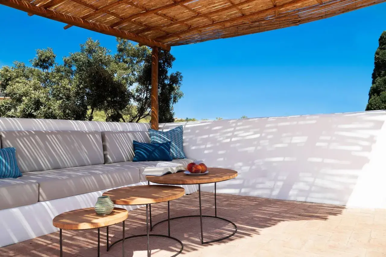 Terrasse ombragée d'un agriturismo en Sardaigne, avec canapé confortable, tables basses en bois et vue sur la végétation luxuriante sous un ciel bleu éclatant.