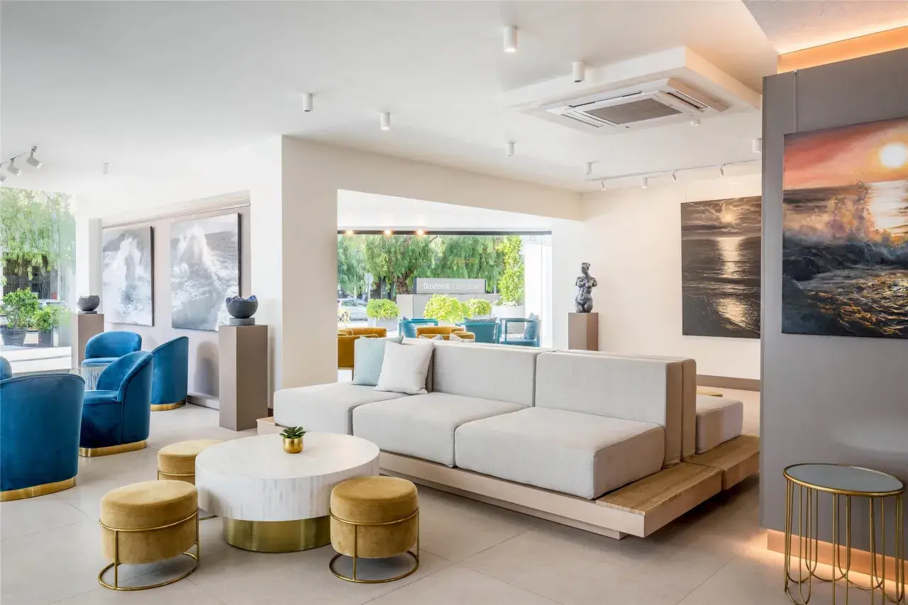 Salón moderno con sofás modulares, sillones azules y arte marino. El cartel "Occidental Puerto Banús" se ve al fondo.