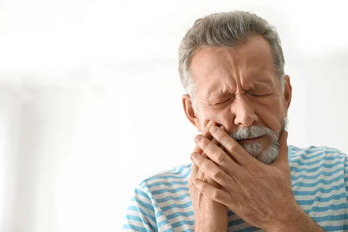 Hombre mayor con los ojos cerrados y la mano en la mejilla, sufriendo de dolor, posiblemente por alveolitis seca.