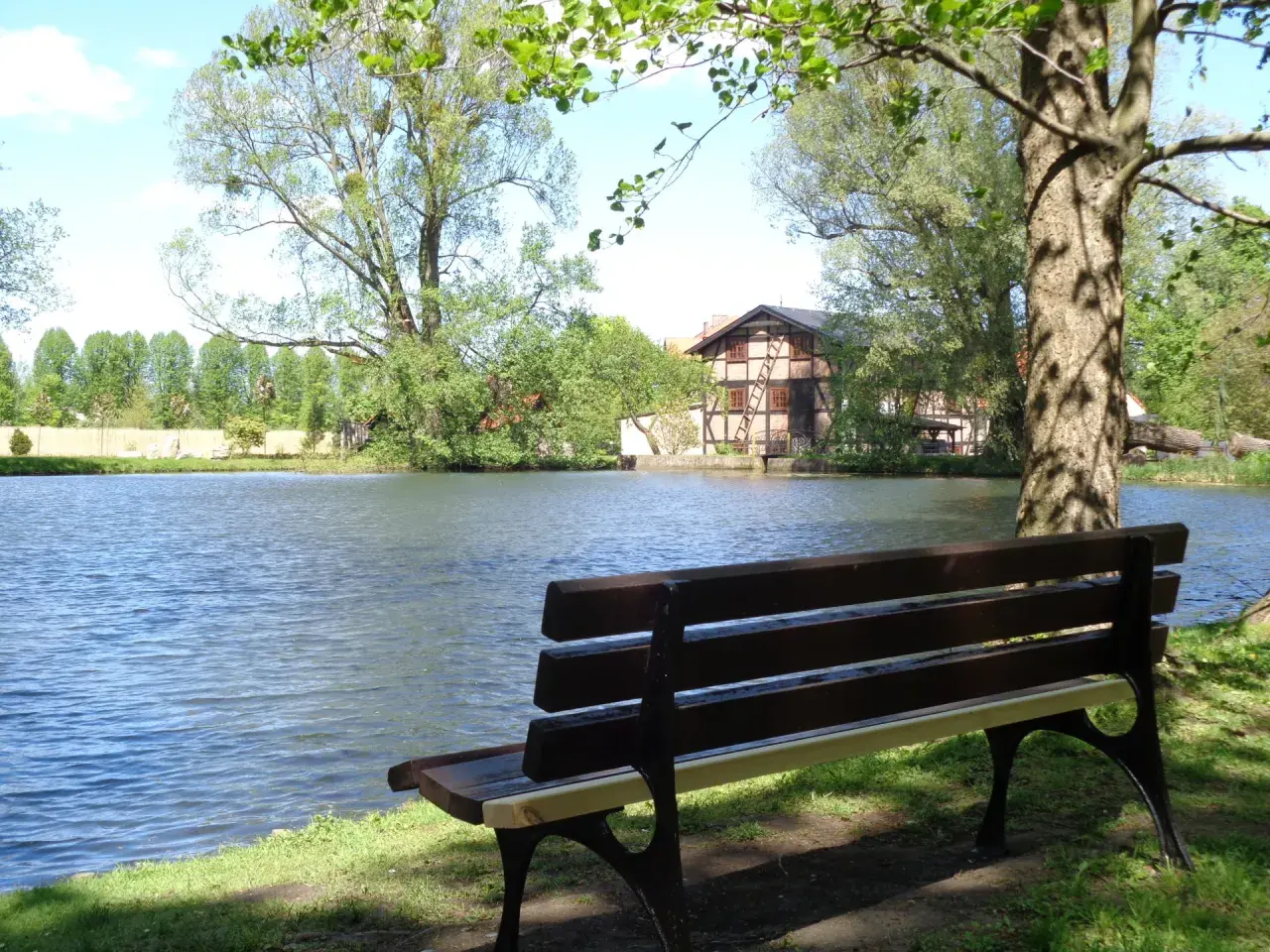 Ławka nad jeziorem, idealne miejsce na chwilę wytchnienia w **dolinie radości**. W tle dom z muru pruskiego i bujna zieleń.