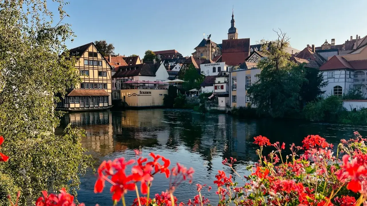 Blick auf Bamberg mit Fachwerkhäusern am Wasser. Rote Geranien blühen im Vordergrund. Entdecken Sie Bamberg Geheimtipps entlang des Flusses.