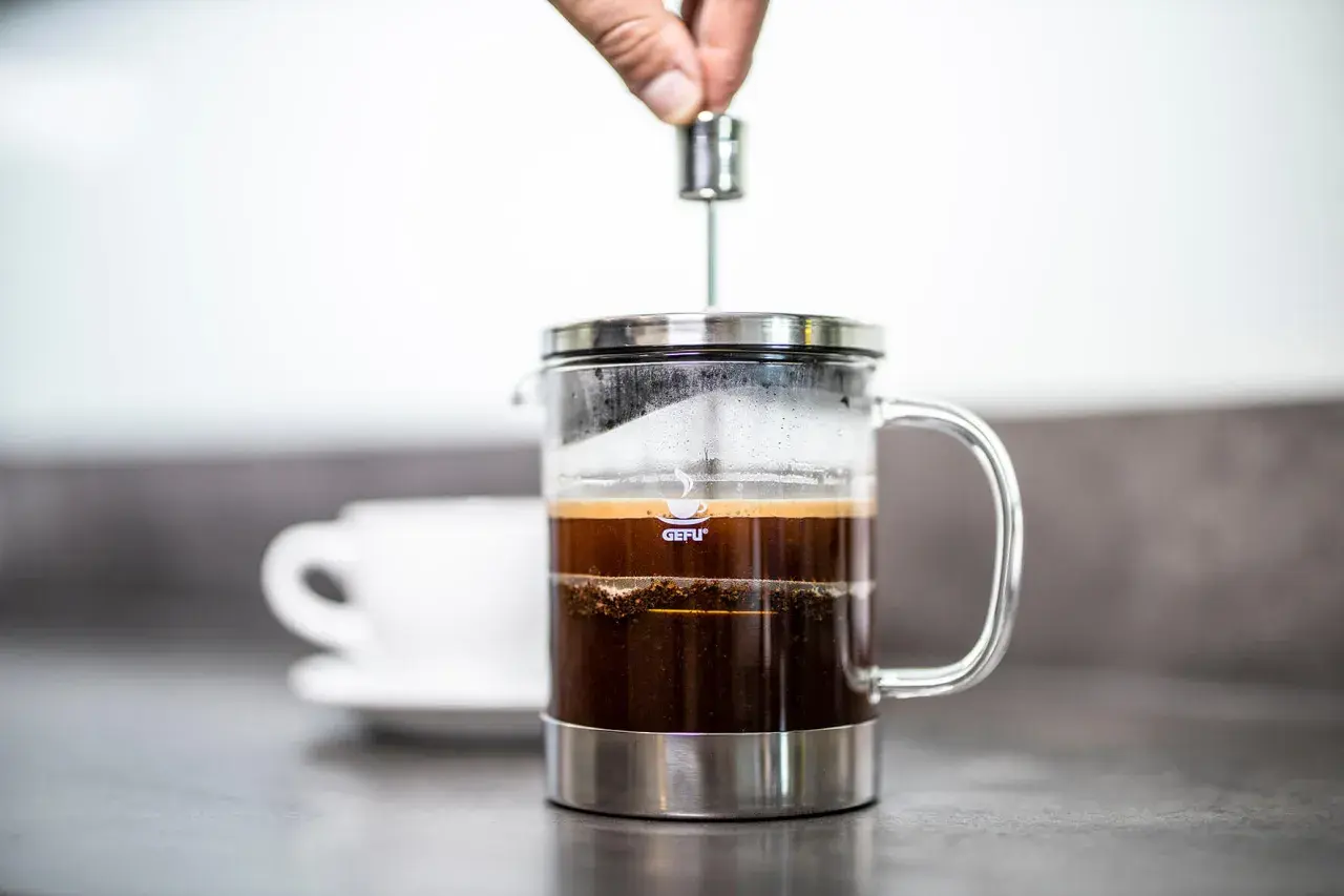 Hand drückt Kolben in French Press, um einen milden Kaffee zuzubereiten. Eine weiße Tasse steht im Hintergrund.