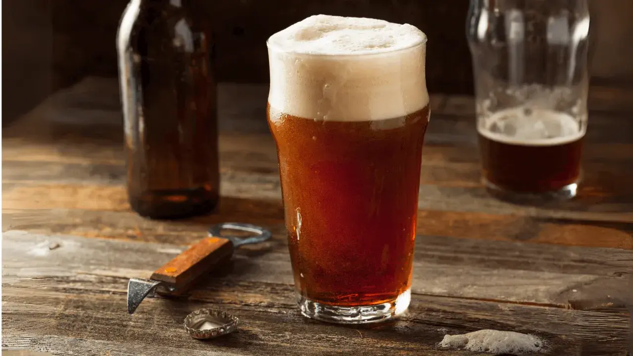 Un bicchiere di birra rossa con schiuma abbondante, accanto a una bottiglia e un apribottiglie. Perfetta per gustare i suoi gradi.