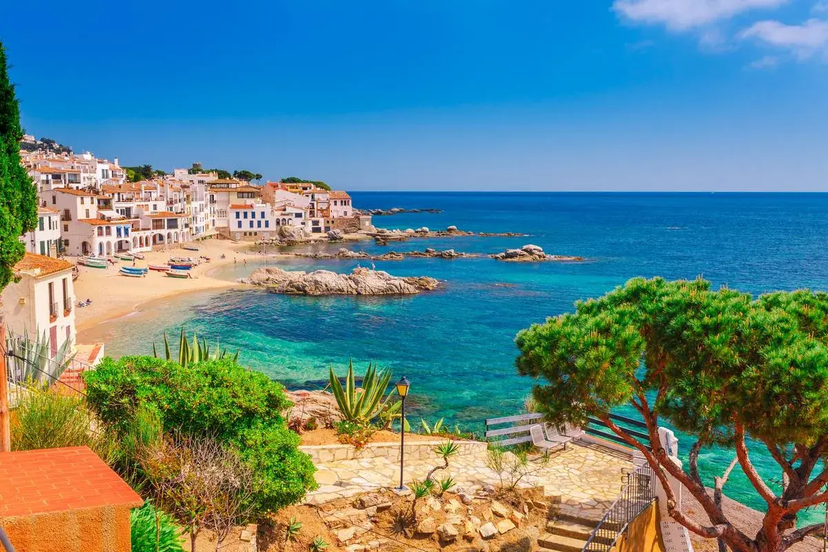 Vue pittoresque d'une crique ensoleillée avec des maisons blanches, des bateaux colorés et des eaux turquoises, parfait pour les activités Costa Brava.