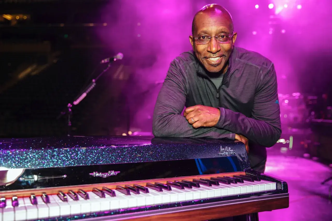 Greg Phillinganes lächelt, lehnt sich an ein glitzerndes Klavier auf einer Bühne im lila Licht.