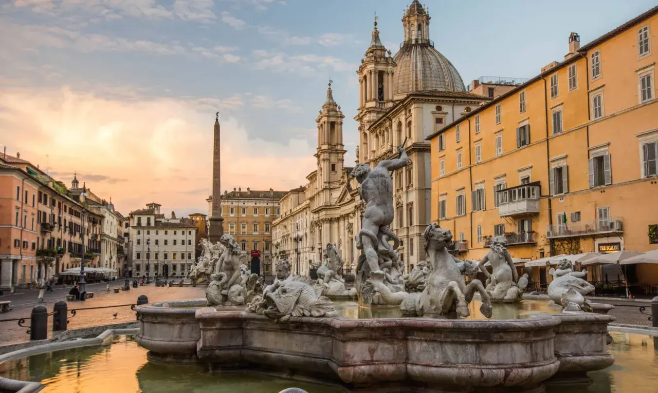 Fontana dei Quattro Fiumi a Piazza Navona, con l'obelisco e la chiesa di Sant'Agnese in Agone sullo sfondo, al tramonto.