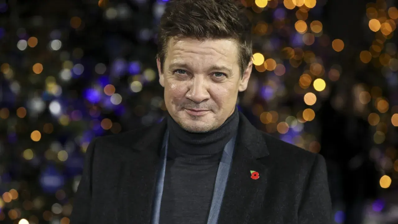 Jeremy Renner, med en vallmo på kavajen, ser allvarlig ut mot en bakgrund av julbelysning. Efter sin svåra olycka är han tillbaka.