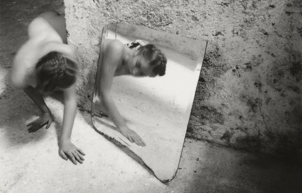 Une femme se contorsionne près d'un miroir brisé, son reflet fragmenté. Une œuvre de Francesca Woodman.