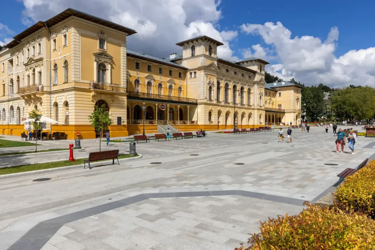 Plac przed zabytkowym sanatorium. Ludzie spacerują, odpoczywają na ławkach. Może to miejsce, gdzie czekają na nowe zasady wyjazdu do sanatorium na NFZ.