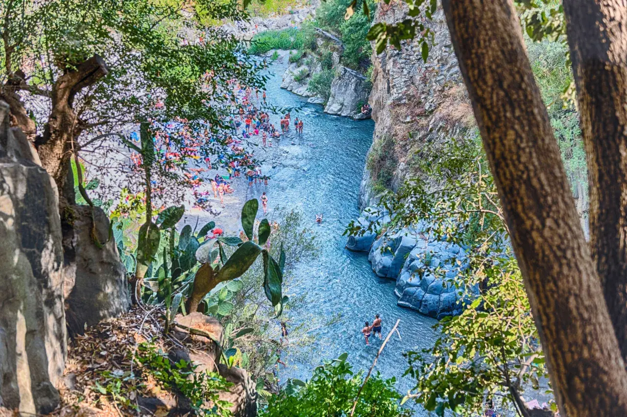 Gente si gode il sole e l'acqua cristallina nella Valle dell'Alcantara, tra rocce vulcaniche e vegetazione rigogliosa.