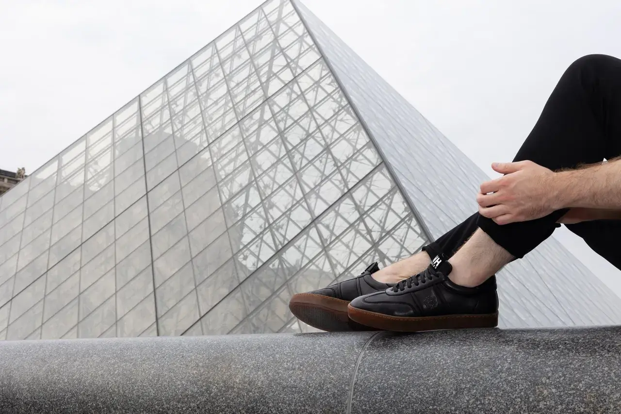 Un homme porte une tenue avec basket noir, assis devant la pyramide du Louvre.