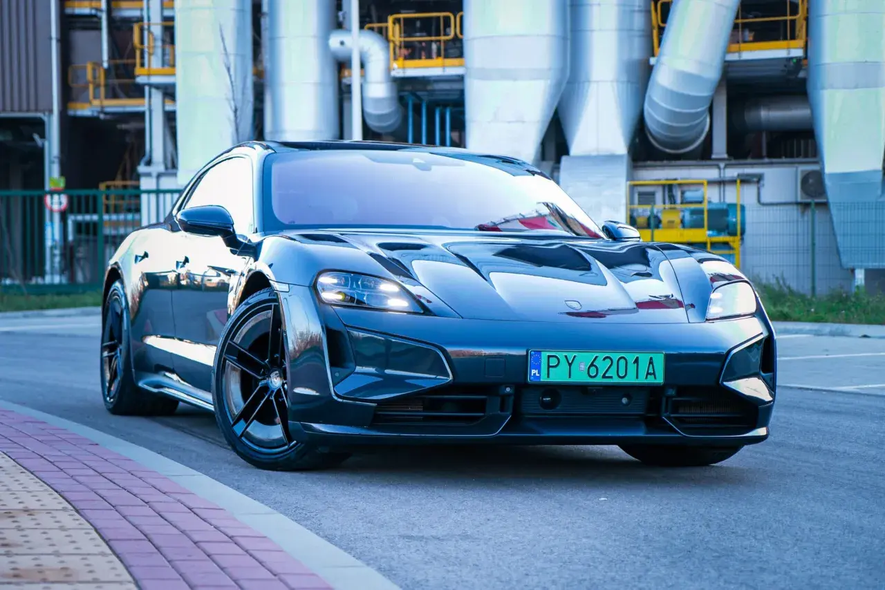 Elegancki Porsche Taycan w ciemnym kolorze, zaparkowany przed industrialnym budynkiem z rurami.