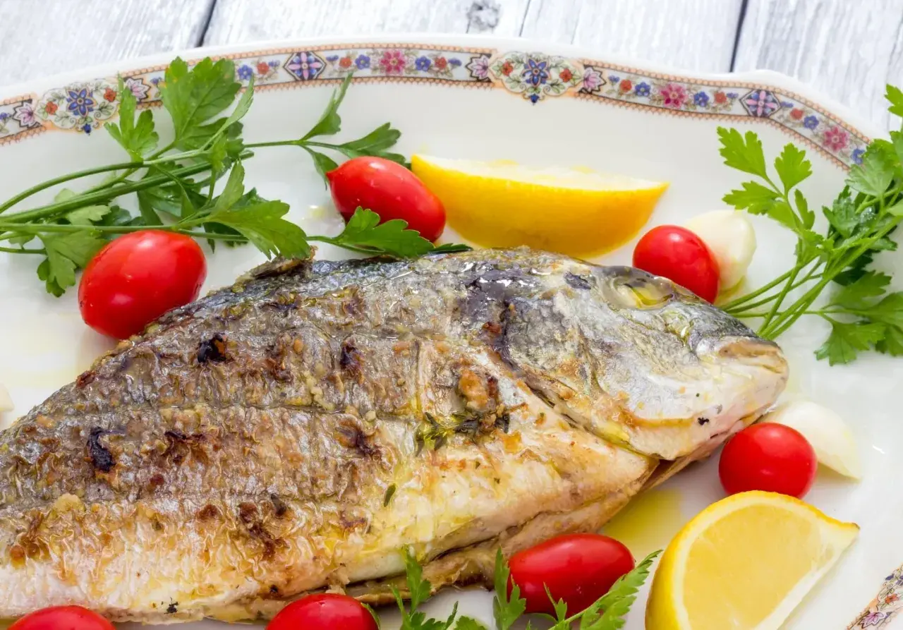 Orata alla griglia dorata e succulenta, guarnita con pomodorini, spicchi d'aglio, limone e prezzemolo fresco.