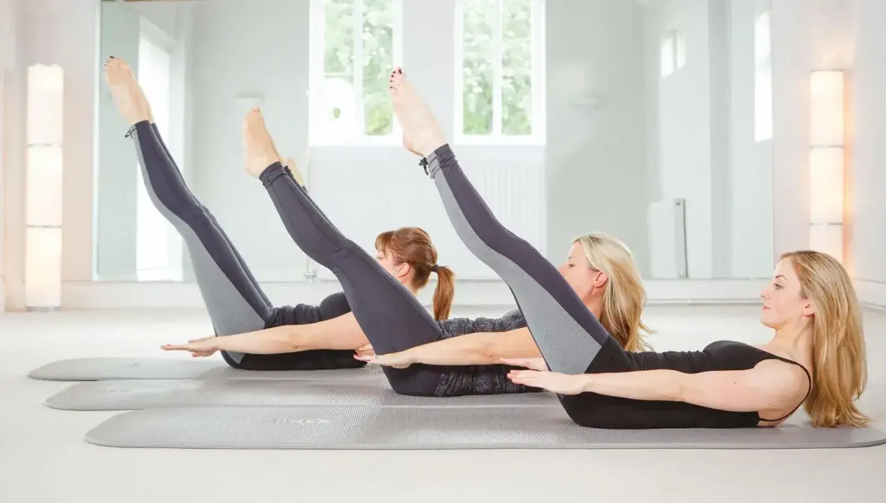 Tre donne eseguono posizioni pilates sul tappetino, sollevando gambe e braccia in un esercizio di core stability.