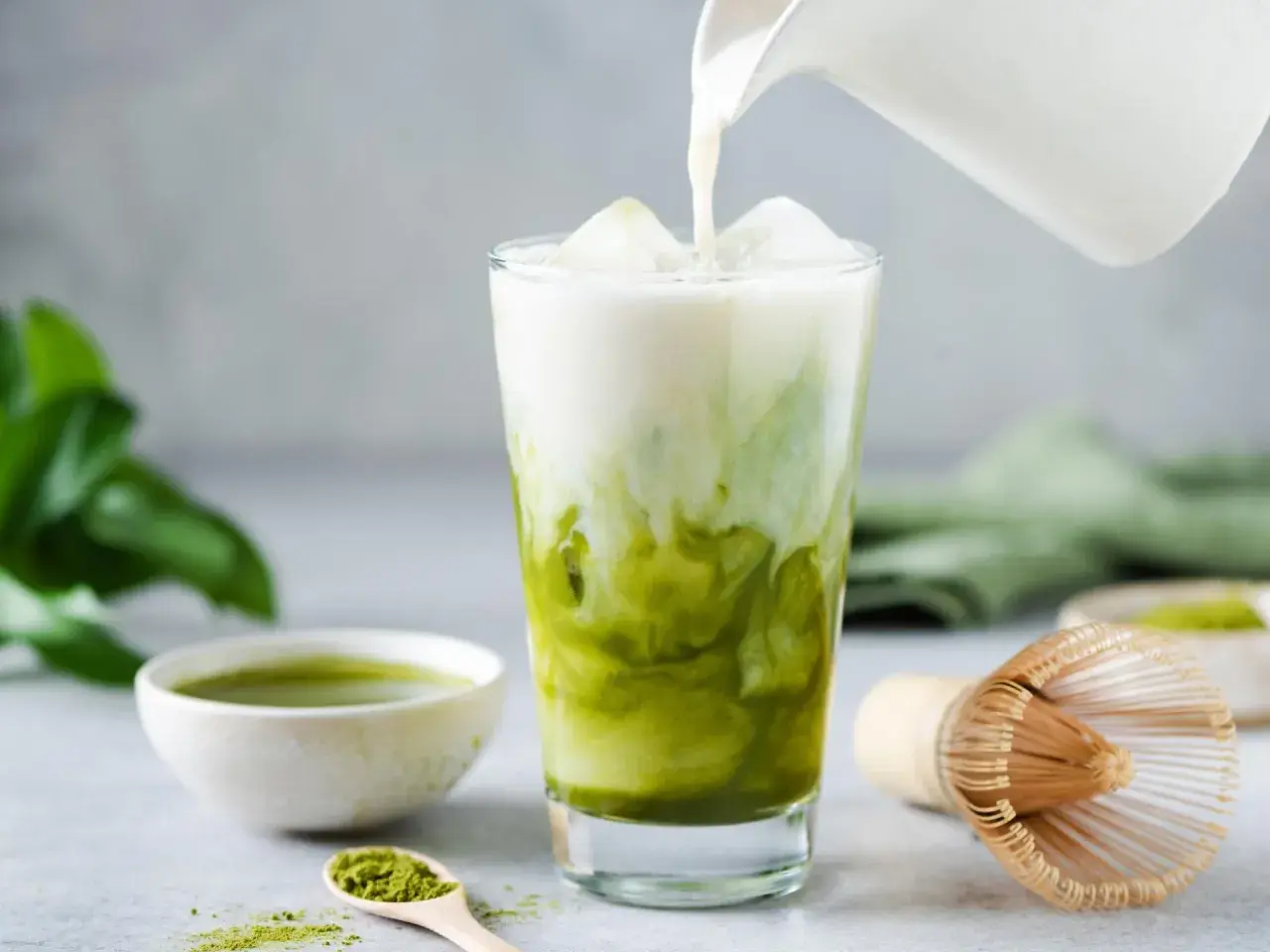 Milch wird in ein Glas mit Eis und Matcha gegossen. Ein Löffel mit Matcha-Pulver und eine Schale mit Matcha-Tee sind daneben zu sehen. Ein traditioneller Matcha-Besen liegt auf dem Tisch.