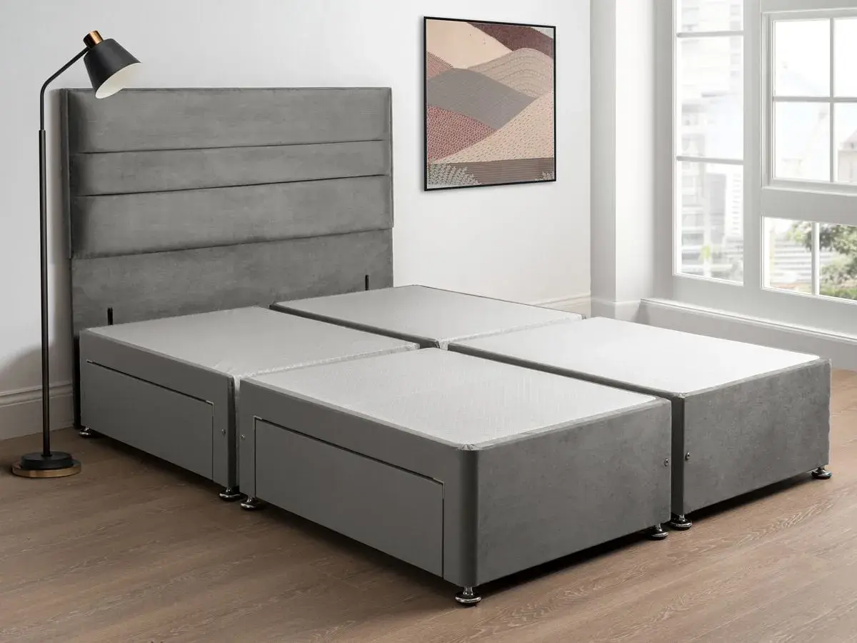 A grey sprung divan base with drawers, paired with a matching headboard, stands in a bright room with a large window.
