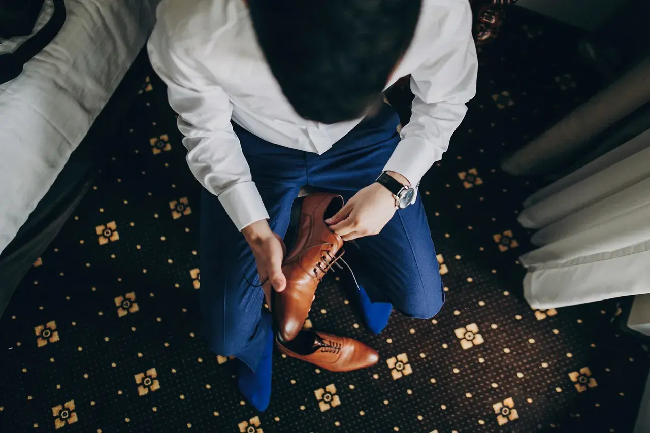 Mężczyzna w niebieskim garniturze i białej koszuli zakłada brązowe, eleganckie buty. Idealne pytanie: jakie buty do niebieskiego garnituru?