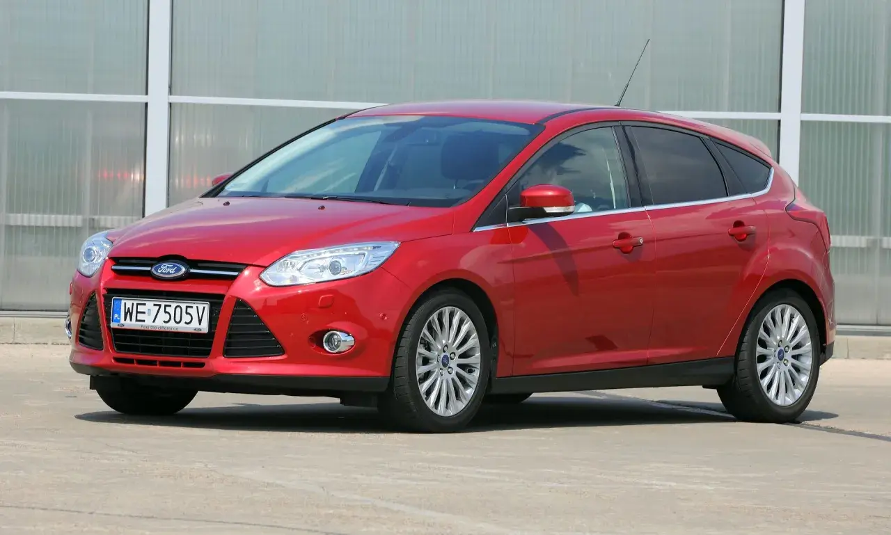 Czerwony Ford Focus z włączonymi światłami mijania, zaparkowany przed nowoczesnym budynkiem.