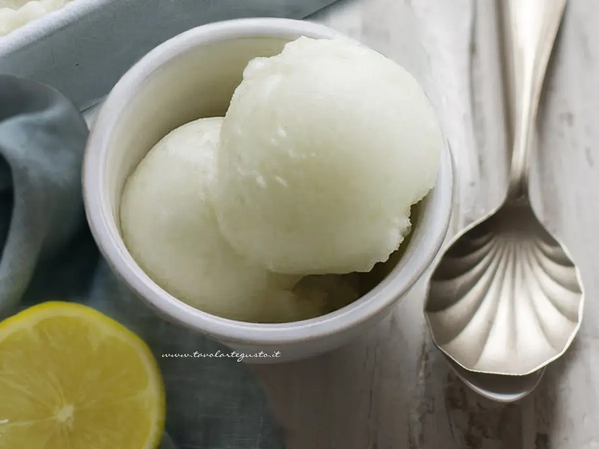 Due palline di sorbetto al limone senza albume in una ciotola bianca, con una fetta di limone e un cucchiaio decorato accanto.