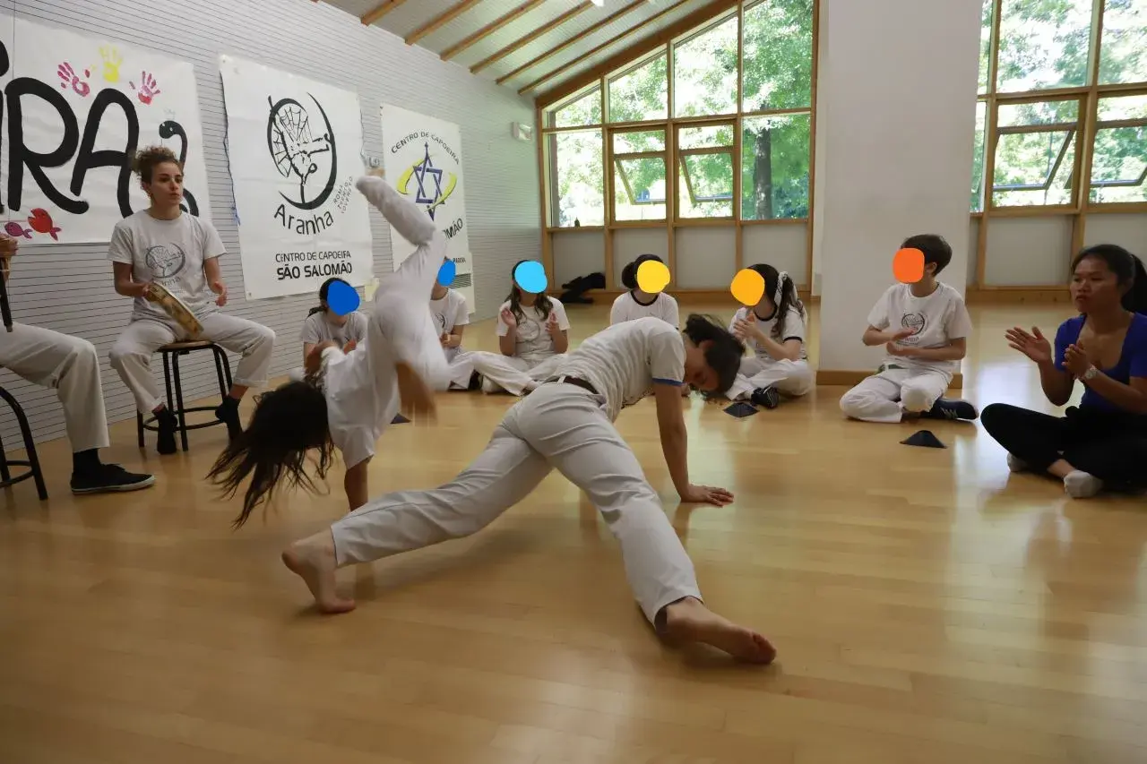 Bambini praticano capoeira in una palestra luminosa. Uno fa una capriola, altri osservano e applaudono.