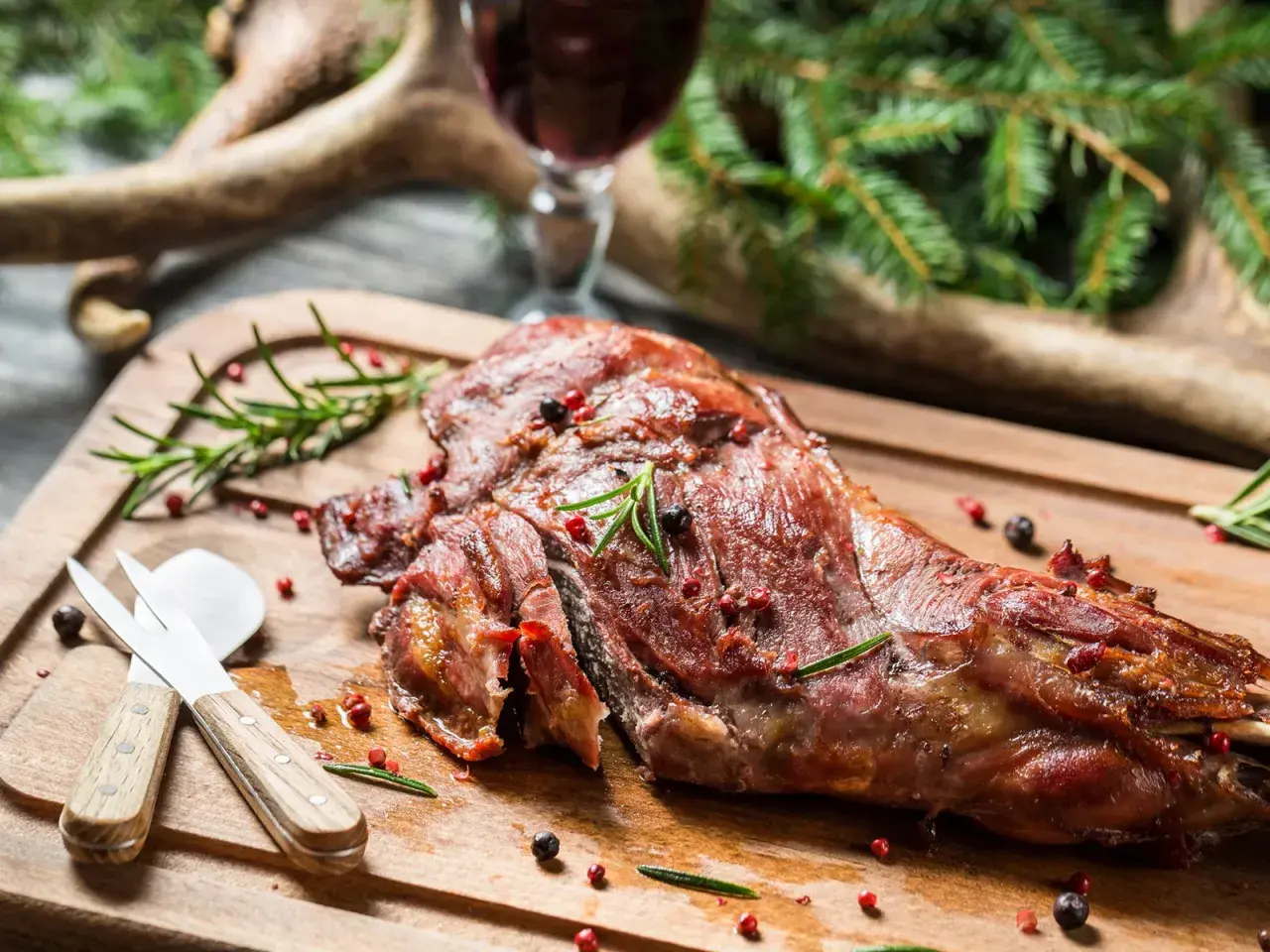 Ein saftiger Rehschlegel, perfekt gegrillt, liegt auf einem Holzbrett. Gewürze und Rosmarin zieren das Fleisch. Ein Glas Rotwein und Hirschgeweih im Hintergrund deuten auf ein festliches rehschlegel rezept hin.