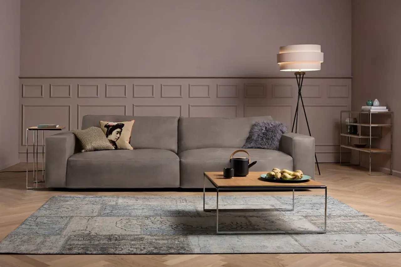 Gemütliches Wohnzimmer mit einem Sofa in Farbe Taupe, einem Couchtisch mit Teekanne und Früchten, sowie einem Teppich.