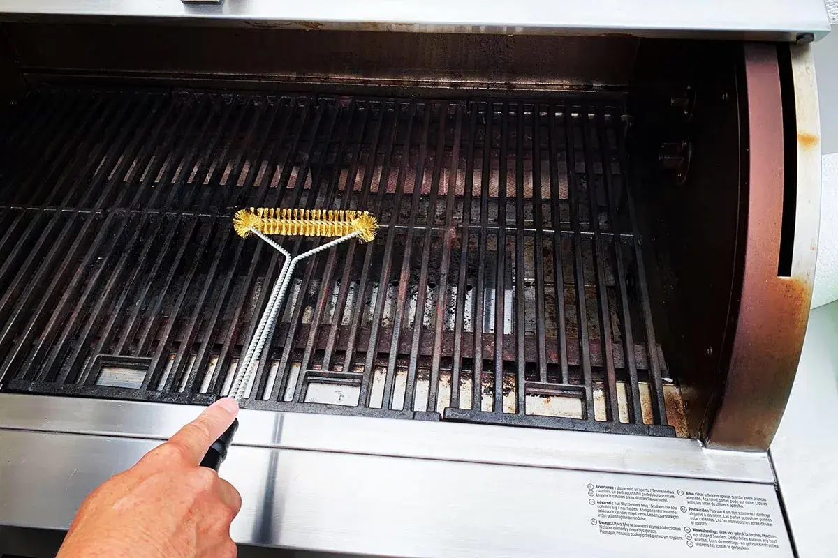 En hand rengör gallret på en gasolgrill under tak med en grillborste.