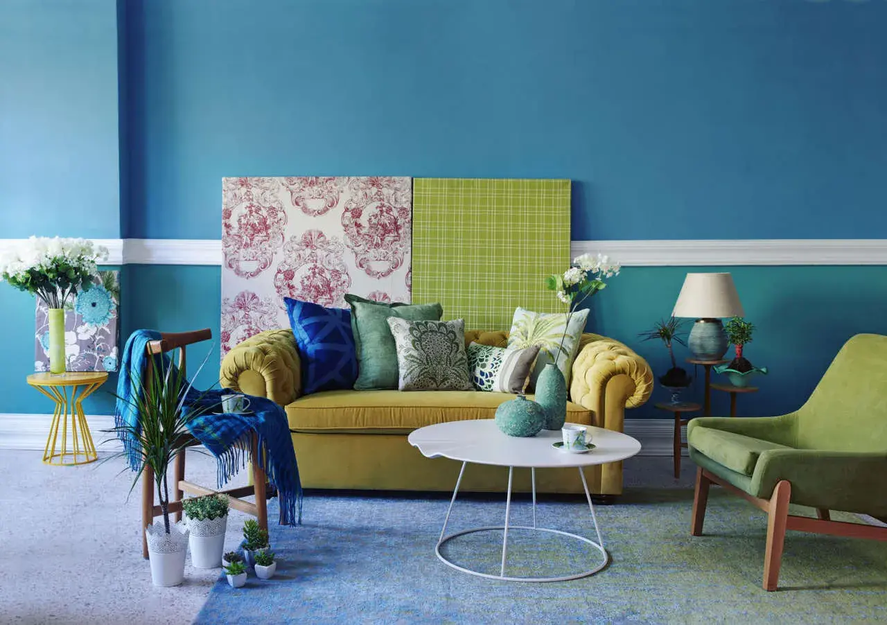Soggiorno con divano giallo, cuscini verdi e blu, e pareti blu. Ottime idee per colorare le pareti di casa con contrasti vivaci.