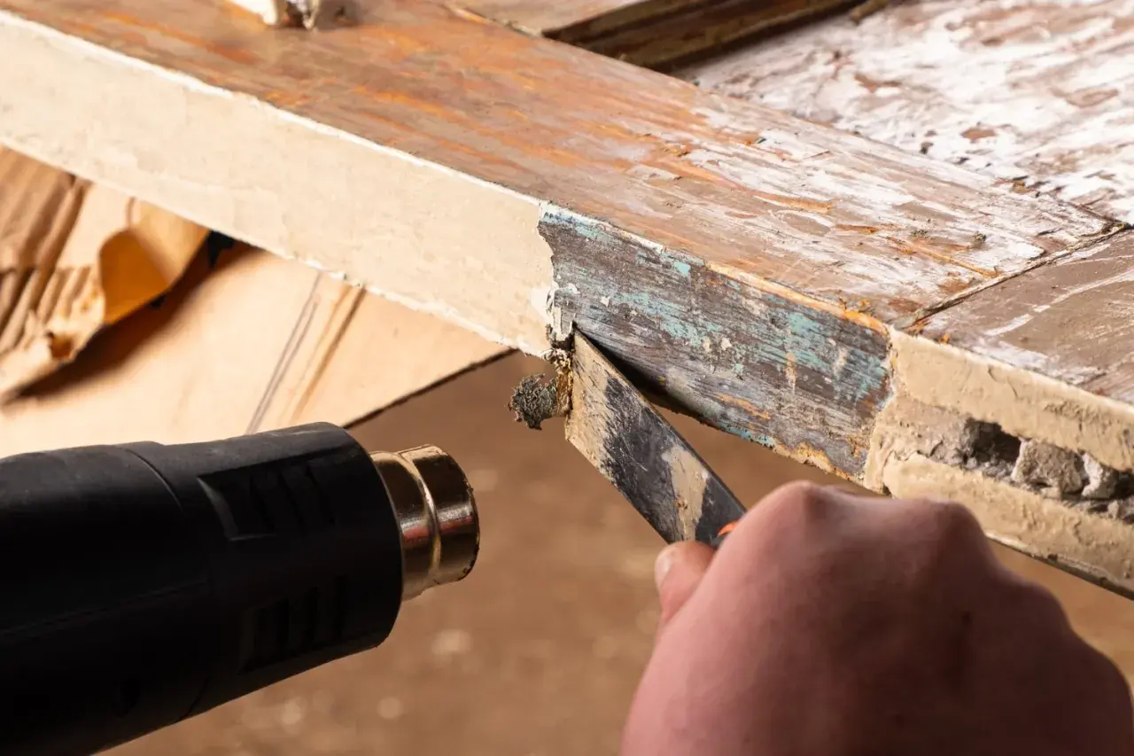 Cómo quitar la pintura de la madera sin lijar: se usa pistola de calor y rasqueta para despegar capas viejas de pintura de un marco de madera.