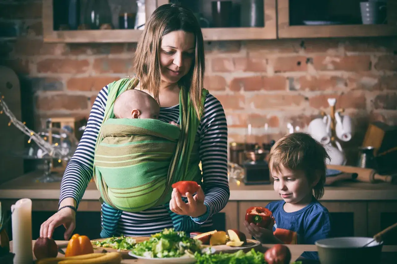 Mama z dzieckiem w chuście i synem przygotowują posiłek. Od kiedy można nosić dziecko w chuście? Od pierwszych dni życia!