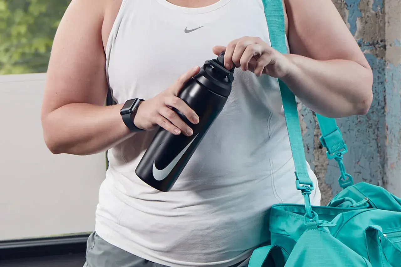 Osoba w białej koszulce sportowej trzyma czarną butelkę Nike. Obok wisi turkusowa torba.