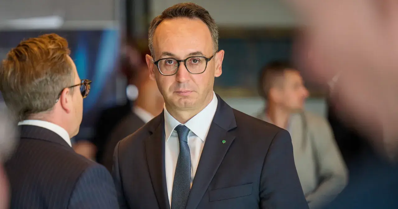 Minister infrastruktury w okularach i garniturze, z zieloną koniczynką na klapie marynarki, rozmawia z kimś na tle innych osób.